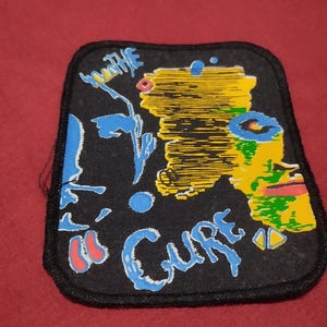 Könnte beinhalten: Ein rechteckiger schwarzer Stoffaufnäher mit gesticktem Rand. Das Design zeigt ein stilisiertes gelb-grünes Gesichtsprofil auf der rechten Seite, blaue abstrakte Formen und den Schriftzug "THE CURE" in blauen Buchstaben auf der linken Seite.