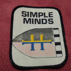 Könnte beinhalten: Ein rechteckiger Stoffaufnäher mit abgerundeten Ecken und einem schwarzen gestickten Rand. Auf dem weißen Hintergrund steht der schwarze Text SIMPLE MINDS über einer schildförmigen Grafik mit einem gelb-blauen Symbol und schwarz-weißen Streifen.