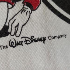 May include: Close-up of white fabric featuring a black and red cartoon character's hand and arm. The Walt Disney Company logo is printed in black, along with the text "The Walt Disney Company" and the number "87".