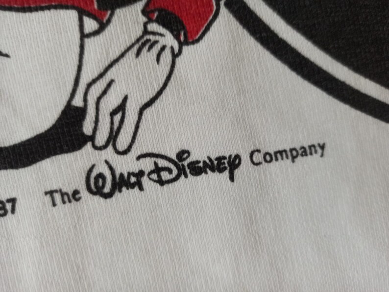 May include: Close-up of white fabric featuring a black and red cartoon character's hand and arm. The Walt Disney Company logo is printed in black, along with the text "The Walt Disney Company" and the number "87".