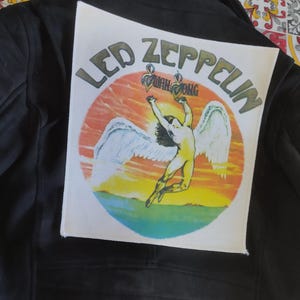 Może przedstawiać: Czarna skórzana kurtka z białą naszywką z logo Led Zeppelin i postacią z skrzydłami na tle zachodu słońca.