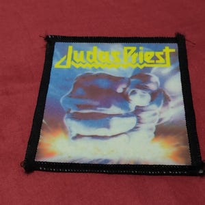 May include: A rectangular fabric patch with a black stitched border. The centre features a graphic of a metallic fist with a bright yellow light effect at the base. The text Judas Priest is printed in yellow lettering across the top of the patch.