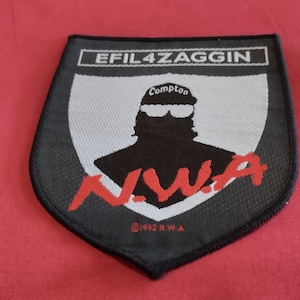 May include: A black, shield-shaped patch with the text "EFIL4ZAGGIN" at the top. The patch features a silhouette of a person wearing sunglasses and the word "Compton". The letters "N.W.A" are in red, and the text "©1992 R.W.A" is at the bottom.