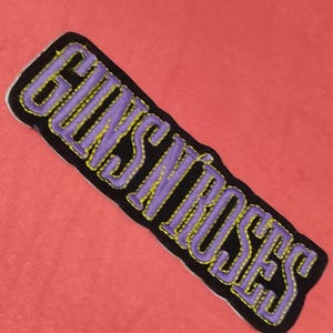 May include: A rectangular patch featuring the words "GUNS N' ROSES" in purple, with yellow stitching, on a black background. The patch is set against a coral-coloured fabric.