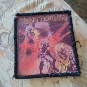May include: A rectangular fabric patch featuring the Iron Maiden logo at the top. The graphic depicts a central figure with long hair and a skeletal face, surrounded by red and orange flames, with a smaller winged creature in the upper left corner.