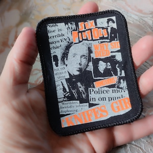 Könnte beinhalten: Ein rechteckiger schwarzer Stoffaufnäher mit einer Collage aus schwarz-weißen Zeitungsausschnitten über die Sex Pistols. Das Design enthält orangefarbenen Text mit Sex Pistols, Why Sid Died und Knifes Gir sowie ein Porträt einer Person.