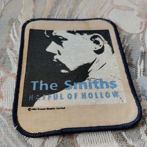 Pode incluir: Um emblema retangular de tecido com uma borda costurada em azul escuro. O design apresenta um perfil em preto e branco de alto contraste de uma pessoa, com o texto "The Smiths" e "HATFUL OF HOLLOW" em azul, e "©1984 Crunch Graphic Limited" abaixo.