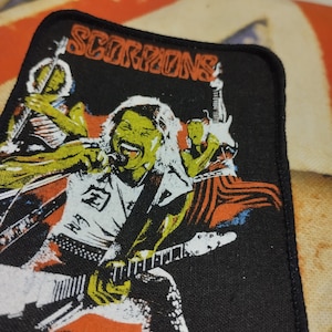 Puede incluir: Parche negro con el nombre de la banda "SCORPIONS" en naranja. El parche muestra a un miembro de la banda tocando la guitarra con un diseño colorido. El parche tiene un borde negro.