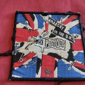 以下が含まれることがあります： ユニオンジャックのデザインの正方形パッチ。テキスト「ANARCHY IN THE U.K.」と「SEX PISTOLS」が特徴です。パッチは安全ピンで飾られ、黒い縁があります。背景は無地の赤色です。