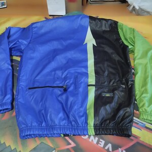 May include: A blue, black, and green windbreaker jacket with a zipper closure and a large white arrow on the back. The jacket has a pocket on the left side and a drawstring at the bottom.