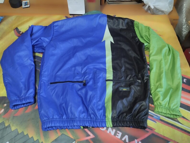 May include: A blue, black, and green windbreaker jacket with a zipper closure and a large white arrow on the back. The jacket has a pocket on the left side and a drawstring at the bottom.