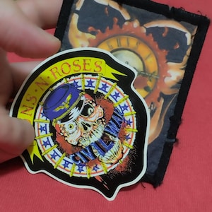 May include: A colourful patch featuring a skull wearing a blue hat and the text "GUNS N' ROSES". The patch has a star and banner design with a black border. A second patch with a clock design is also visible.