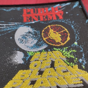 Peut inclure: Un écusson noir avec le texte rouge "PUBLIC ENEMY" au-dessus d'un graphique de planète et d'un emblème jaune et rouge. Les mots "FEAR OF A BLACK PLANET" sont en jaune en dessous.