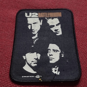 Puede incluir: Un parche rectangular de tela negra con un gráfico serigrafiado en blanco de la banda U2. El texto U2 RATTLE AND HUM está impreso en la parte superior. El borde inferior incluye el texto 1989 NOT US LTD con un pequeño logotipo. Los bordes están cosidos.