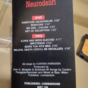 以下が含まれることがあります： 白黒のカセットテープケースで、タイトルは「Neurodeliri」。曲名とその長さが分単位と秒単位で表示されています。ケースには、バンド名、プロデューサー、出版社、そして「Metalmaster」のラベルも記載されています。
