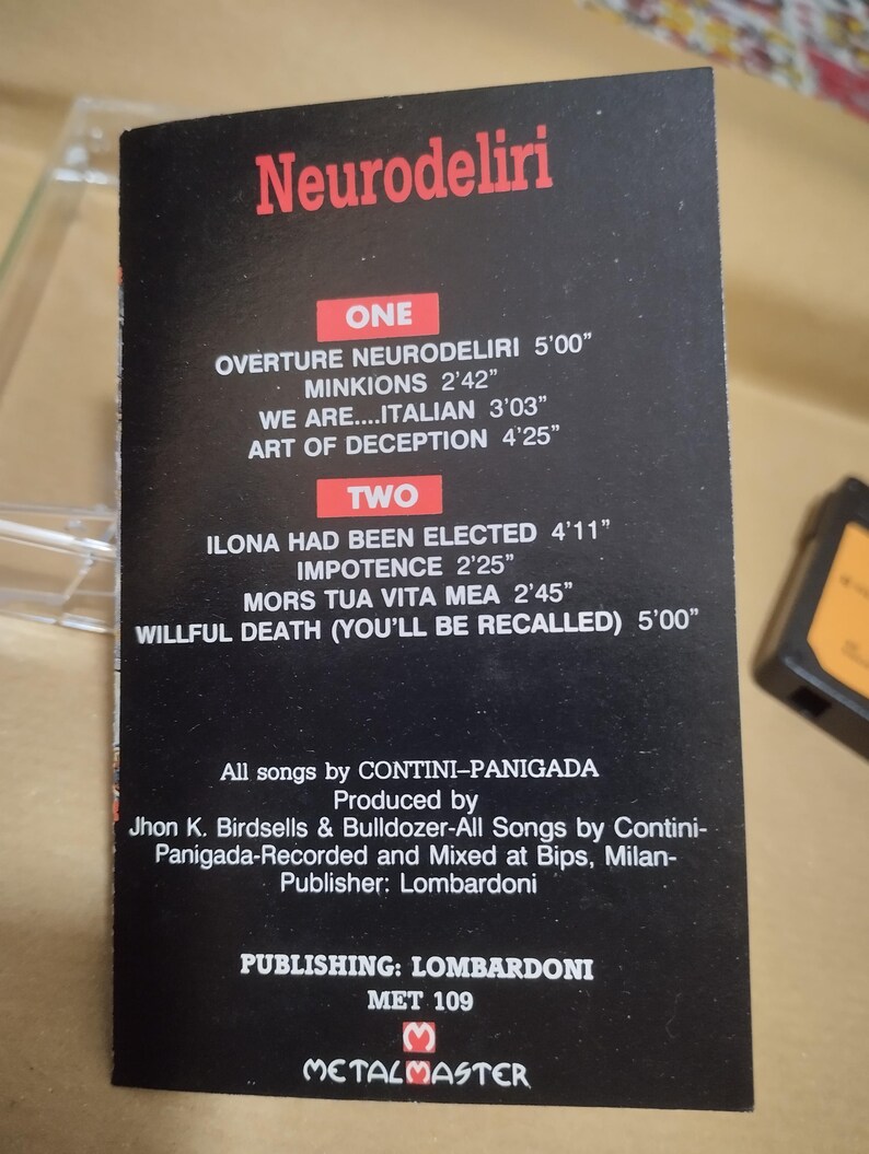 以下が含まれることがあります： 白黒のカセットテープケースで、タイトルは「Neurodeliri」。曲名とその長さが分単位と秒単位で表示されています。ケースには、バンド名、プロデューサー、出版社、そして「Metalmaster」のラベルも記載されています。