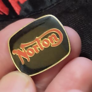 May include: A gold-coloured pin with a black background and the word "Norton" in red lettering.