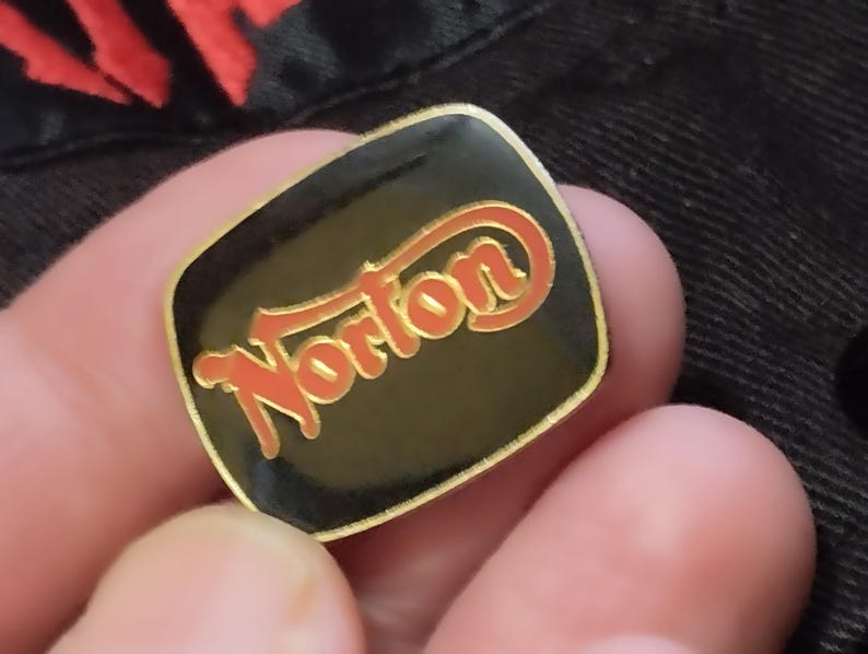 May include: A gold-coloured pin with a black background and the word "Norton" in red lettering.