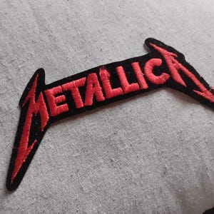 May include: An embroidered fabric patch featuring the word METALLICA in red thread with a black border. The lettering is stylized with elongated, sharp points on the first M and the final A, creating a distinct, angular logo design.