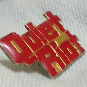 May include: A red and gold metal pin with the word "Quiet Riot" in gold lettering.