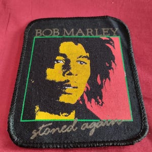 Puede incluir: Parche rectangular negro con un retrato de Bob Marley en amarillo y negro, sobre un fondo rojo y verde. El texto "BOB MARLEY" está en la parte superior y "stoned again" en la inferior.