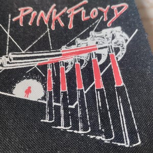Op de afbeelding: Een zwarte stoffen patch met de tekst PINK FLOYD in rode letters aan de bovenkant. De afbeelding toont een reeks rode en witte hamers op een zwarte achtergrond, met een klein rood figuurtje dat voor een witte cirkel staat.