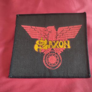 Può includere: Una toppa quadrata in tessuto nero con una grafica di un'aquila rossa con le ali spiegate. La parola SAXON è stampata in giallo al centro dell'aquila e un emblema circolare rosso con un motivo geometrico è posizionato sotto il testo.