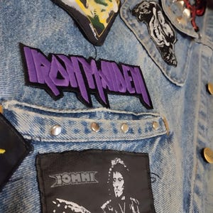 Op de afbeelding: Close-up van een spijkerjasje versierd met patches en metalen studs. De jas heeft een grote paarse "IRON MAIDEN" patch, samen met andere band patches en zilveren studs. De jas heeft een klassiek ontwerp met knopen.