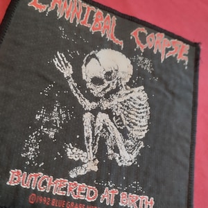 Könnte beinhalten: Schwarzer quadratischer Aufnäher mit dem roten Schriftzug "CANNIBAL CORPSE" über einer weißen Skelettillustration. Darunter steht der Text "BUTCHERED AT BIRTH" in Rot. Der Aufnäher hat den Text "©1992 BLUE GRAPE MERCHANDISING CO."