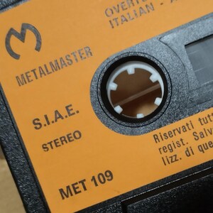 以下が含まれることがあります： オレンジ色のカセットテープのクローズアップ。テキストは「METALMASTER」、「S.I.A.E.」、「STEREO」、「MET 109」、「Riservati tutti i diritti regist. Salvo autori lizz. di questo prodotto」です。
