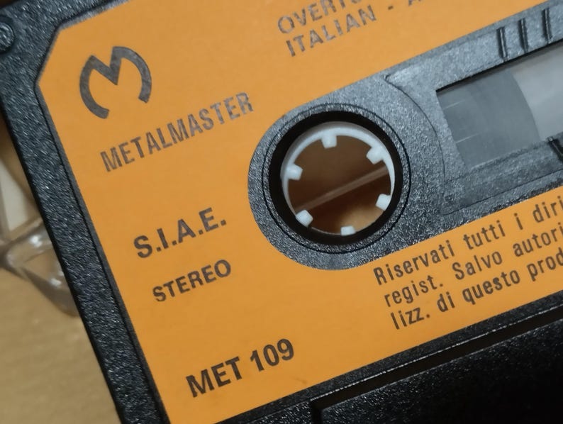 以下が含まれることがあります： オレンジ色のカセットテープのクローズアップ。テキストは「METALMASTER」、「S.I.A.E.」、「STEREO」、「MET 109」、「Riservati tutti i diritti regist. Salvo autori lizz. di questo prodotto」です。
