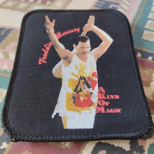 Könnte beinhalten: Ein rechteckiger schwarzer Stoffaufnäher mit genähtem Rand, der Freddie Mercury in einem weißen Tanktop mit erhobenen Armen zeigt. Der Schriftzug Freddie Mercury erscheint in roter Schreibschrift und A KIND OF MAGIC ist in Rot gedruckt.