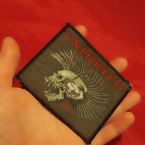 Pode incluir: Um patch quadrado com a palavra "EXPLOITED" em letras vermelhas. O patch apresenta uma caveira com moicano e linhas radiantes em um fundo cinza. O patch é segurado em uma mão.