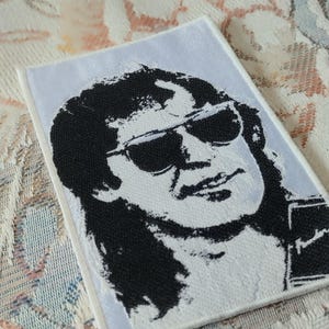 May include: A rectangular embroidered patch featuring a high-contrast black and white portrait of a person with long hair wearing dark sunglasses. The design includes a small rectangular graphic in the bottom right corner with the text Fender.
