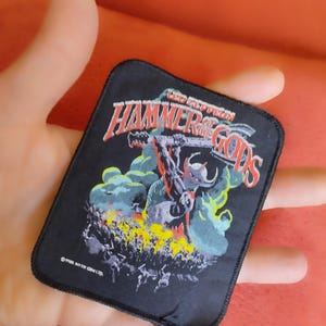 Pode incluir: Um emblema retangular de tecido preto com uma ilustração gráfica de uma figura com chifres segurando um martelo. O texto diz LED ZEPPELIN e HAMMER OF THE GODS em letras vermelhas e brancas. O design inclui efeitos de fumaça amarela e verde.
