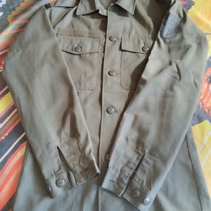 May include: A green long-sleeved shirt with a button-up front and two pockets. The shirt has a patch on the right shoulder.