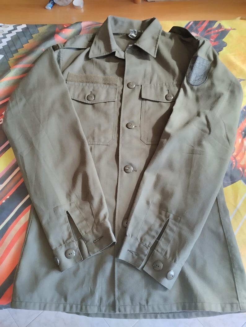 May include: A green long-sleeved shirt with a button-up front and two pockets. The shirt has a patch on the right shoulder.
