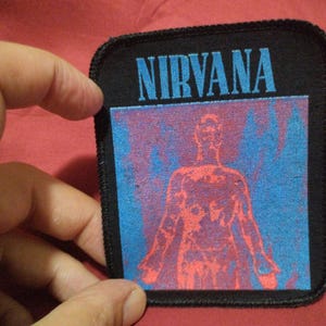 May include: A rectangular black fabric patch with a stitched border. The top features the word NIRVANA in blue text. Below the text is a square graphic depicting a human figure in red against a blue background, resembling a thermal image.