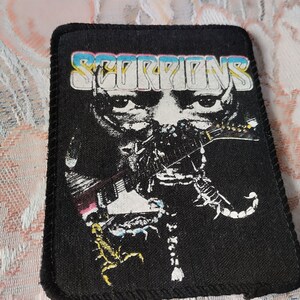 Könnte beinhalten: Ein rechteckiger schwarzer Stoffaufnäher mit dem Wort SCORPIONS in einer stilisierten weißen Schrift mit blauen und rosa Umrandungen. Die Grafik zeigt ein Gesicht mit einer Gitarre und zwei Skorpionen in Weiß- und Gelbtönen auf schwarzem Grund.