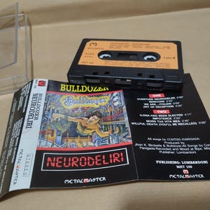 以下が含まれることがあります： 白いラベルに「Bulldozer Neurodeliri」とバンドのロゴが書かれた黒いカセットテープ。カセットテープは透明なプラスチックケースに入っています。ケースの裏面には、バンド名、アルバムタイトル、トラックリストが書かれた白いラベルがあります。