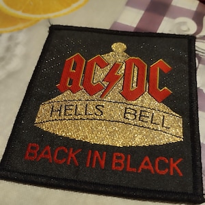 Könnte beinhalten: Schwarzer quadratischer Aufnäher mit dem Text "AC/DC" in Rot, einem goldenen Glitzerdesign und den Worten "HELLS BELL" und "BACK IN BLACK" in Rot. Der Aufnäher liegt auf einer weißen Oberfläche.