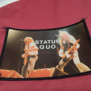 May include: A rectangular fabric patch featuring a colour photograph of the band Status Quo performing on stage. The image shows two musicians playing electric guitars with the white text STATUS QUO printed in the centre. The patch has a black border.
