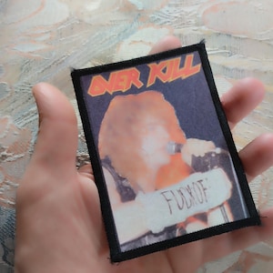 Peut inclure: Un écusson rectangulaire en tissu avec une bordure noire cousue, présentant l'image d'une personne chantant dans un microphone. Le texte OVER KILL apparaît en orange en haut, et le texte FUCK OF est imprimé en noir sur une bannière blanche.