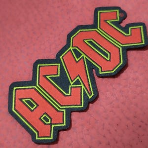 May include: An embroidered patch with the AC/DC logo. The letters are red with a yellow outline and a black border. The lightning bolt is red. The patch is on a red surface.