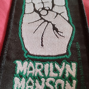 Può includere: Toppa nera con un gesto della mano bianca, con indice e mignolo estesi, bordata di verde. Il nome "MARILYN MANSON" è in bianco con un bordo verde sotto il simbolo della mano. La toppa ha un bordo nero.