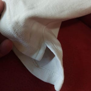 May include: Close-up of white fabric, possibly from a t-shirt or other garment. The image focuses on the texture and stitching of the material. The fabric appears to be soft and lightweight.