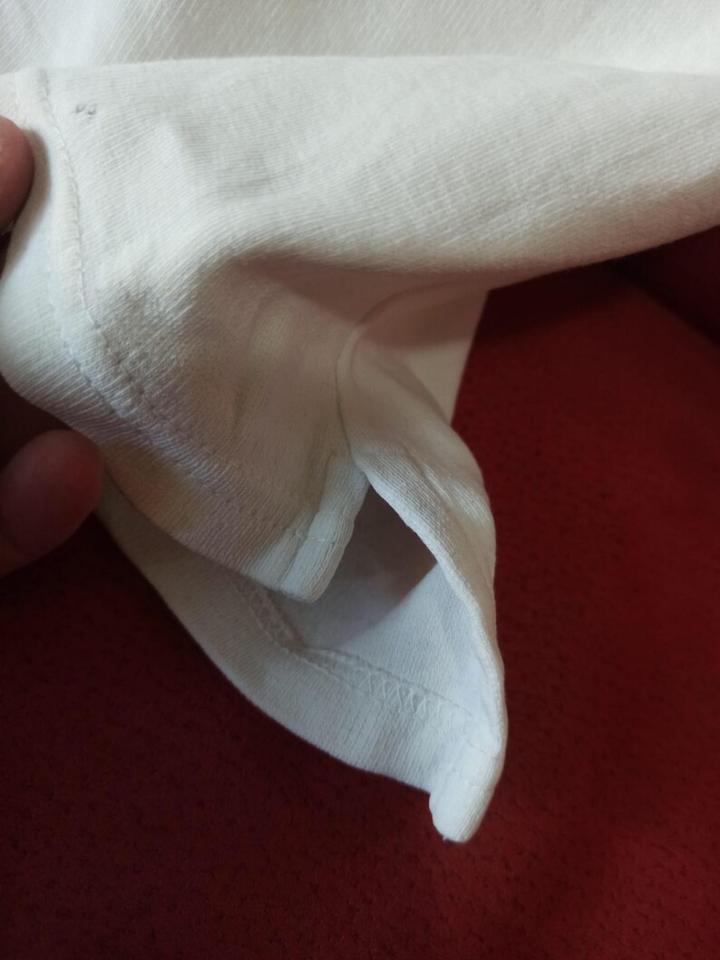 May include: Close-up of white fabric, possibly from a t-shirt or other garment. The image focuses on the texture and stitching of the material. The fabric appears to be soft and lightweight.