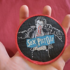 May include: Round black patch with a red border featuring a punk rock graphic. The image depicts a figure with spiky hair and blood, with the words "Sex Pistols" on a blue banner. The patch is likely for clothing or accessories.