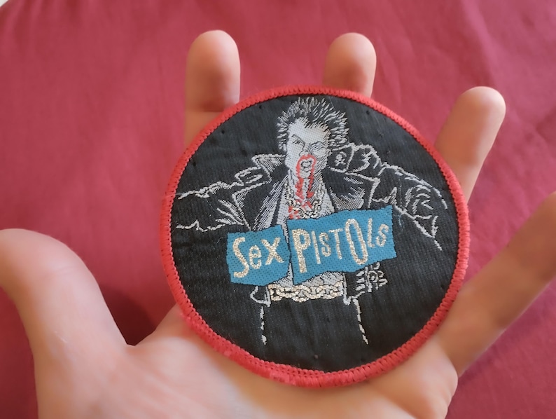 May include: Round black patch with a red border featuring a punk rock graphic. The image depicts a figure with spiky hair and blood, with the words "Sex Pistols" on a blue banner. The patch is likely for clothing or accessories.