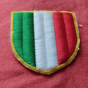 Peut inclure: Un écusson brodé en forme de bouclier présentant des bandes verticales vertes, blanches et rouges. L'écusson est souligné par une bordure jaune vif et se compose de coutures denses et texturées sur toute la surface du motif.
